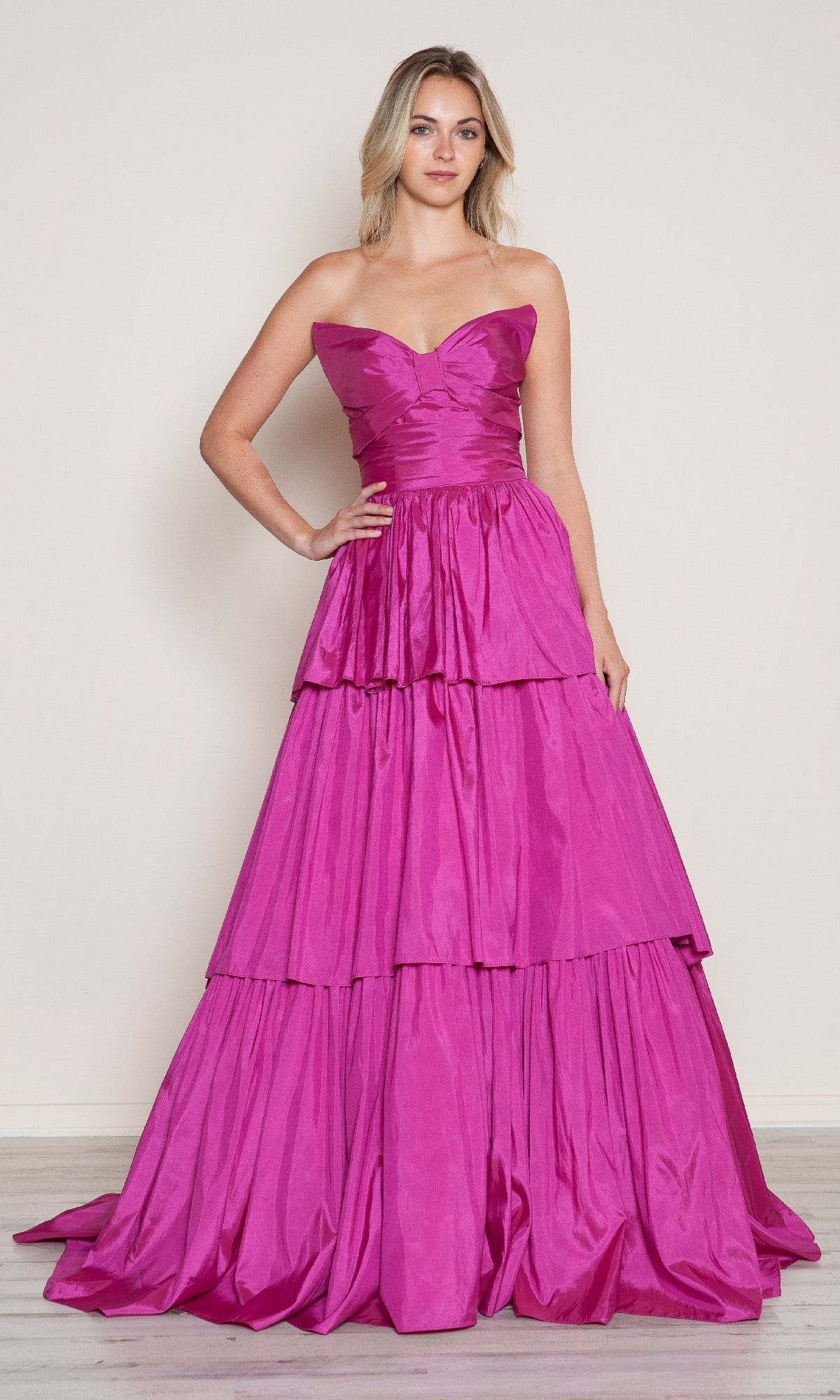 A woman stands against a light-colored wall wearing the Long Prom Dress: Poly USA 9694, a strapless gown with tiered, bright pink layers.